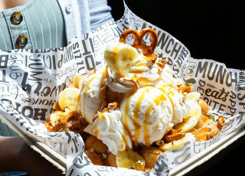 A large dessert with ice cream, whipped cream, bananas, pretzels, and caramel drizzle in a paper tray.