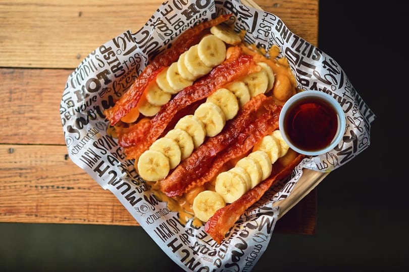 Crispy bacon strips and banana slices on a waffle, with a side of syrup.