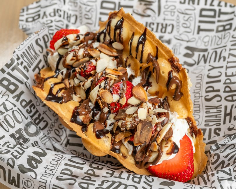 Bubble waffle with strawberries, bananas, whipped cream, chocolate drizzle, and almonds in a paper cone.