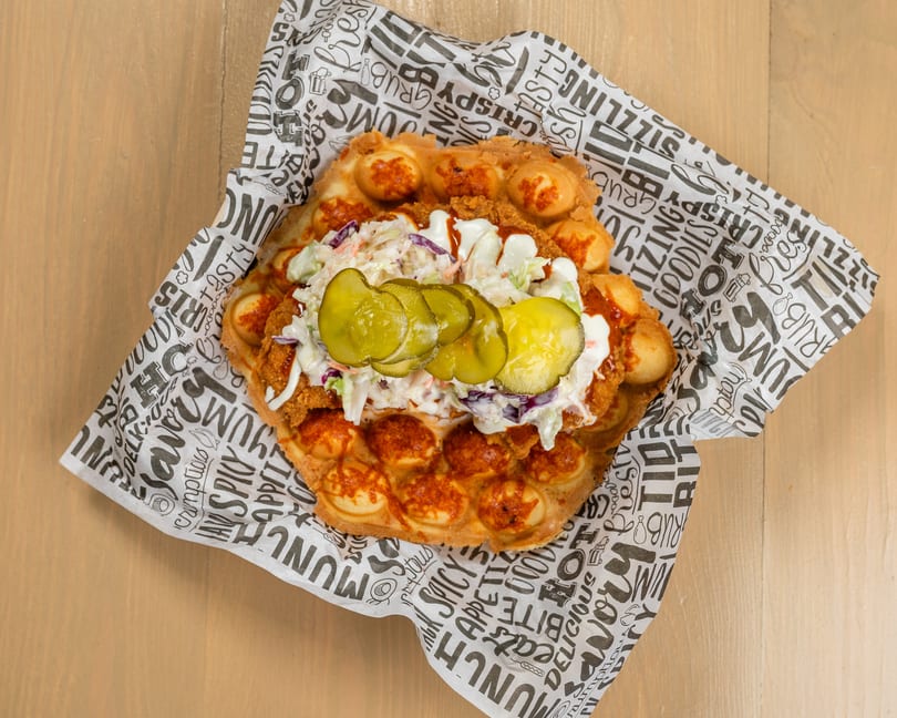 Crispy fried chicken, coleslaw, and pickles on a bubble waffle, served on patterned paper.