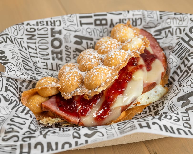 Bubble waffle sandwich with ham, melted cheese, and raspberry jam, dusted with powdered sugar.